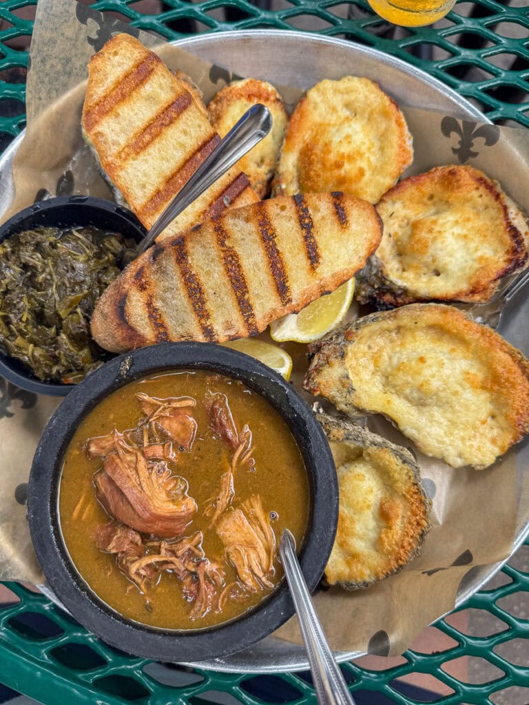 A tray with grilled bread slices, baked oysters, a black bowl of greens, and gumbo with shredded meat sits on brown paper atop a green metal table—perfect fuel before exploring New Orleans attractions with your Go City New Orleans Pass.
