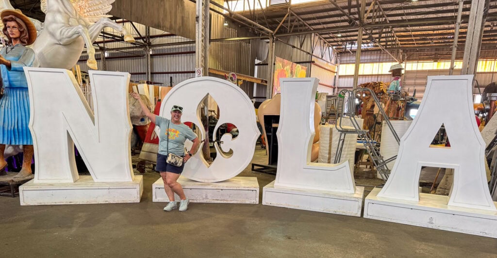 A person poses and smiles in front of large white NOLA letters inside a warehouse, surrounded by Mardi Gras parade floats—one of the top New Orleans attractions you can explore to save money in New Orleans with a Go City New Orleans Pass.