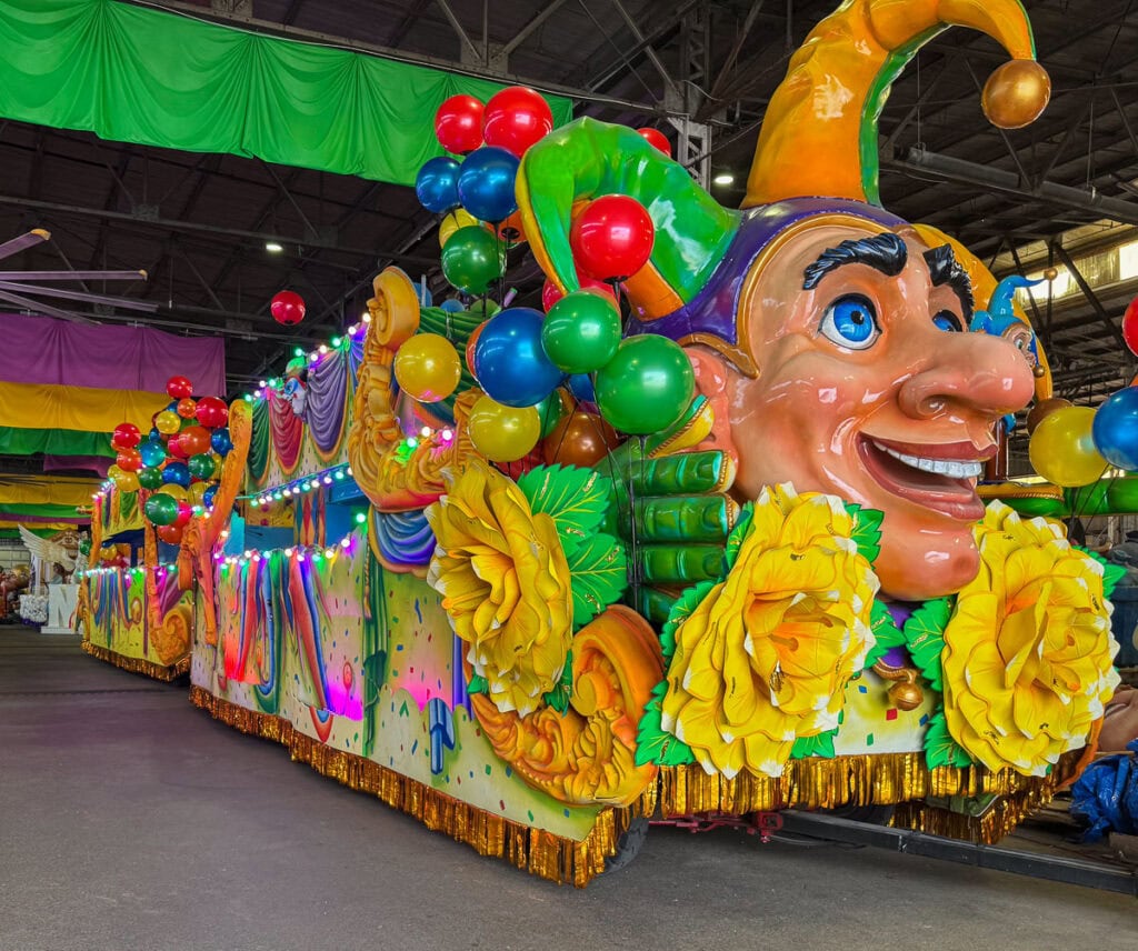 A vibrant Mardi Gras parade float decorated with colorful balloons, yellow flowers, and a large jester head with a yellow hat shines indoors—perfect for photos while you save money on top New Orleans attractions with your Go City New Orleans Pass.