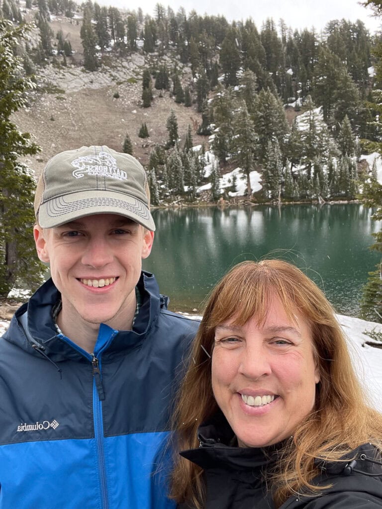 A smiling woman and young man pose for a selfie outdoors in front of a scenic lake surrounded by evergreen trees and snow-dusted hills in one of America's underrated national parks.