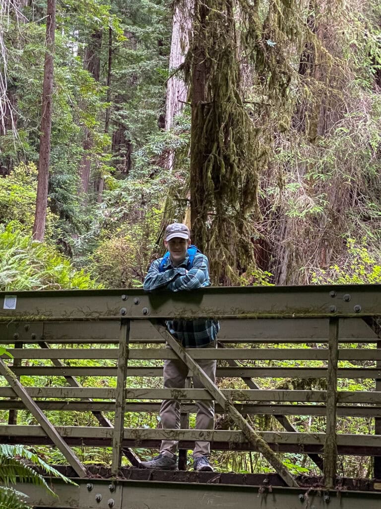 A person wearing a hat and backpack stands on a metal bridge in a lush forest, surrounded by tall trees, ferns, and hanging moss in one of America’s underrated national parks.