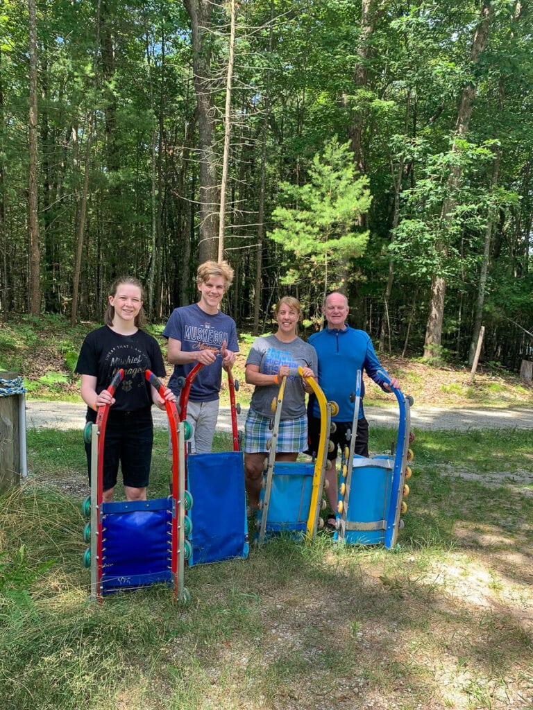 Four people stand outdoors on grass in front of trees, each holding a beach cart with blue seats and yellow or red frames, smiling at the camera on a sunny day—showing how easy it is to fit vacations into a busy family schedule.