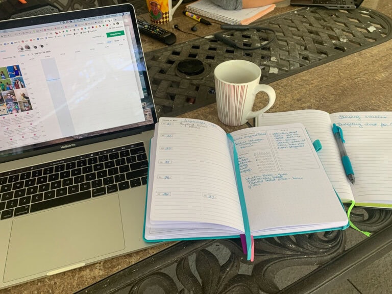 A laptop next to two open notebooks, a pen, and a striped coffee mug on a table. The handwritten notes and dates hint at how to plan a family vacation planning or family travel, while the laptop screen displays a social media page.
