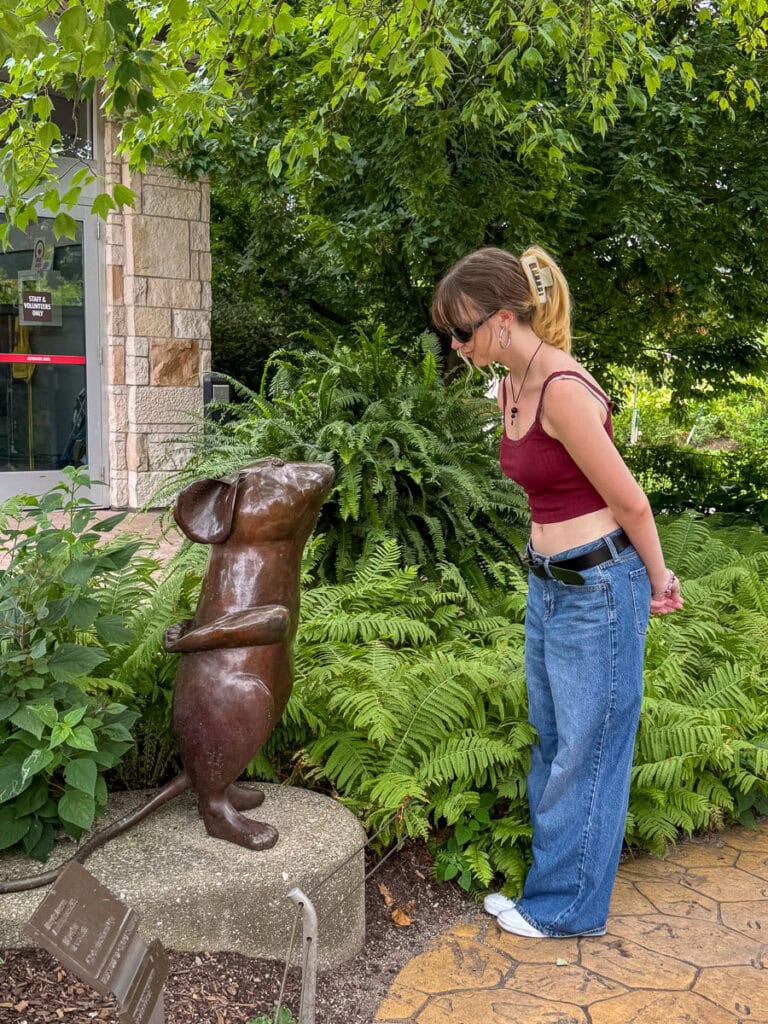 A woman in a red top and blue jeans leans forward, looking at a bronze mouse statue surrounded by green plants—one of the fun things to do in Grand Rapids with teens in a beautiful garden setting.