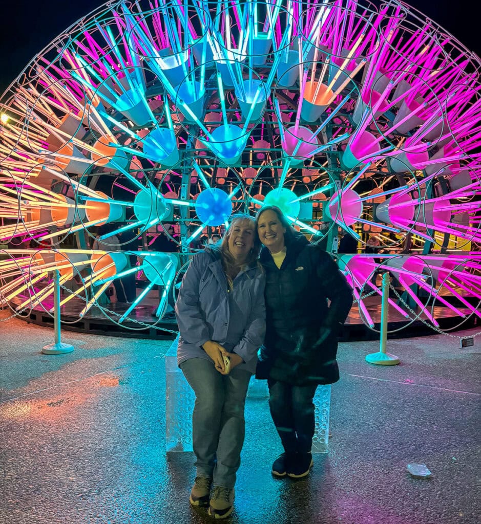 Two women smile while posing together at night in front of a large, colorful, circular neon light installation—one of the top things to do in Grand Rapids with teens. They're wearing jackets and jeans, standing on wet pavement.