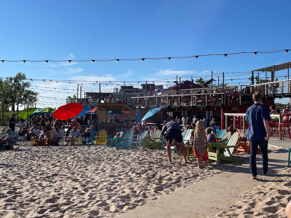 People gather in a sandy outdoor area with colorful chairs, umbrellas, and string lights overhead—one of the fun things to do in Michigan with teens. Buildings and trees sit in the background under a clear blue sky.