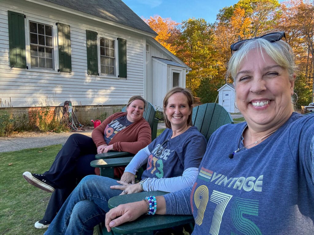 Three smiling women sit on green lawn chairs outside a white house, wearing shirts with "Vintage" and different years. The background captures fall in Michigan with vibrant autumn trees and colorful leaves under a clear sky.