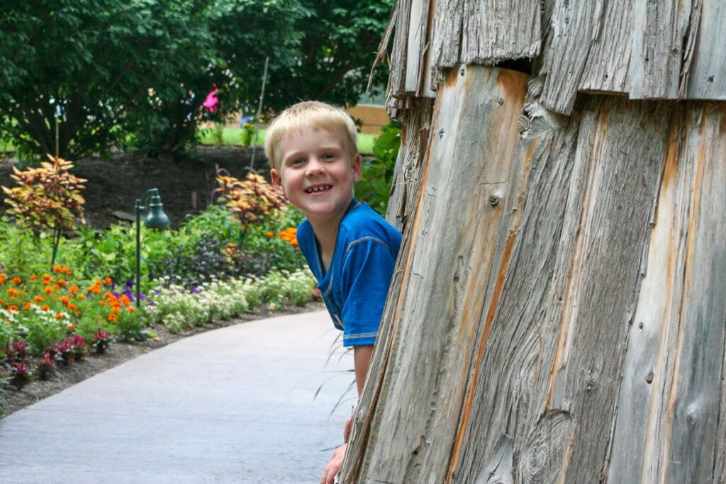 A young boy in a blue shirt smiles while peeking out from behind a rustic wooden structure along a garden path lined with colorful flowers and lush greenery—the scene captures the charm of spring, the Best Time to Visit Grand Rapids.