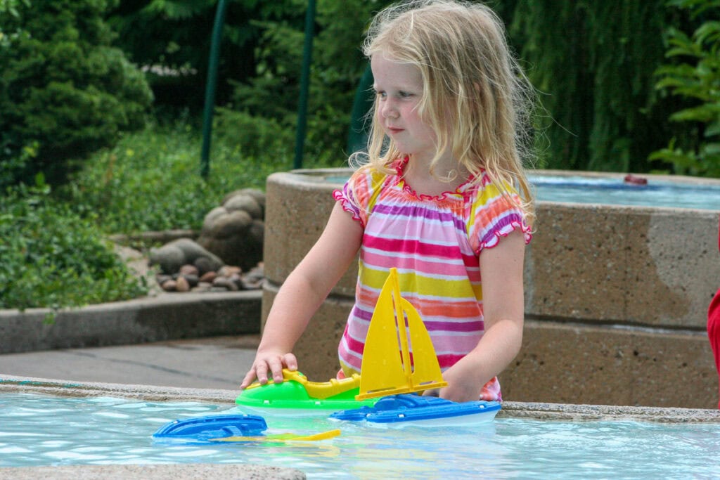 A young girl with blonde hair and a striped dress plays with colorful toy sailboats in a shallow outdoor water feature, surrounded by greenery—a perfect scene during the best time to visit Grand Rapids.