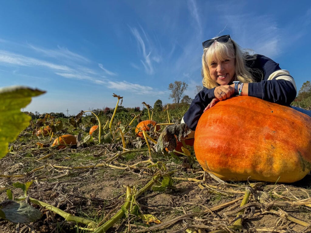 A smiling woman with blonde hair leans on a large orange pumpkin in a pumpkin patch under a blue sky, surrounded by green vines and scattered pumpkins—capturing the charm of autumn, the best time to visit Grand Rapids.