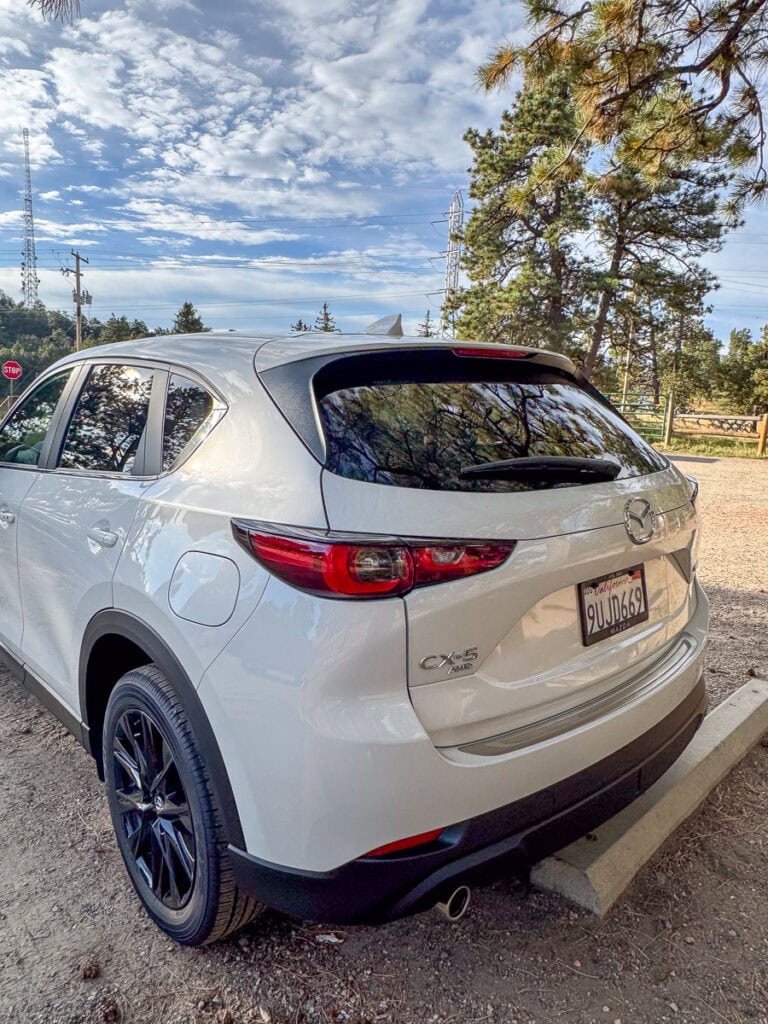 A white Mazda CX-5 SUV, great for road trips, is parked outdoors near trees under a partly cloudy sky, with a California license plate visible and a wooden fence in the background.