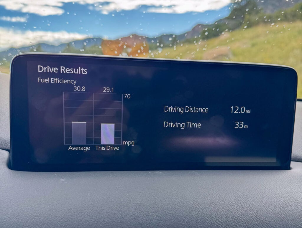 A car dashboard screen displays drive results: fuel efficiency average is 30.8 mpg, this drive is 29.1 mpg. Driving distance is 12.0 miles; driving time is 33 minutes. Rainy landscape is visible—makes you wonder, is the Mazda CX-5 good for road trips?.