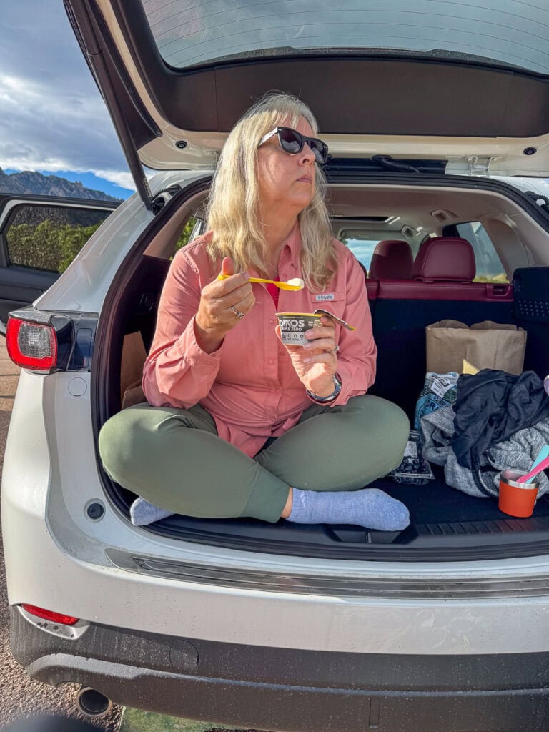 A woman with long blonde hair and sunglasses sits cross-legged in the open trunk of an SUV, eating yogurt with a spoon. The car, possibly a Mazda CX-5—ideal for road trips—is parked outdoors with mountains in the background.