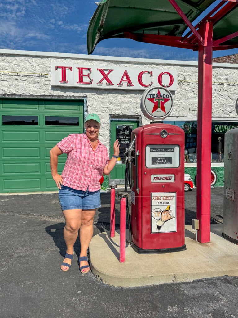 A smiling woman in a pink plaid shirt and denim shorts poses next to a vintage red Texaco gas pump—one of the charming historical landmarks in Kansas—in front of a white building with a green garage door and a large "TEXACO" sign.