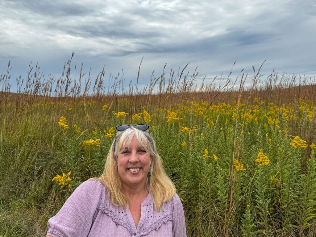 A woman with blonde hair, sunglasses on her head, and a light purple top smiles in front of tall grass and yellow wildflowers under a cloudy sky, capturing the natural beauty near historic sites in Kansas.