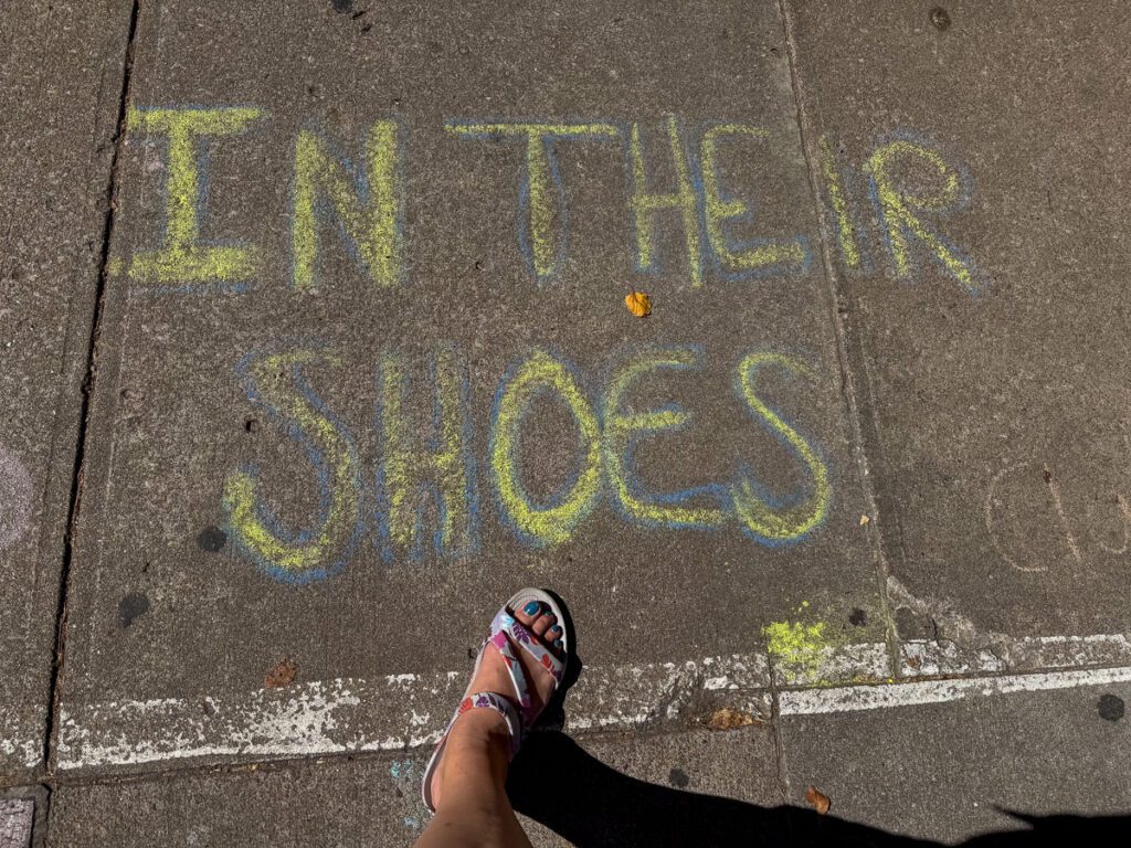 A person's foot in a sandal stands near a sidewalk chalk message in yellow and blue that reads, "IN THEIR SHOES," capturing a moment while visiting with teens in Lawrence, Kansas.