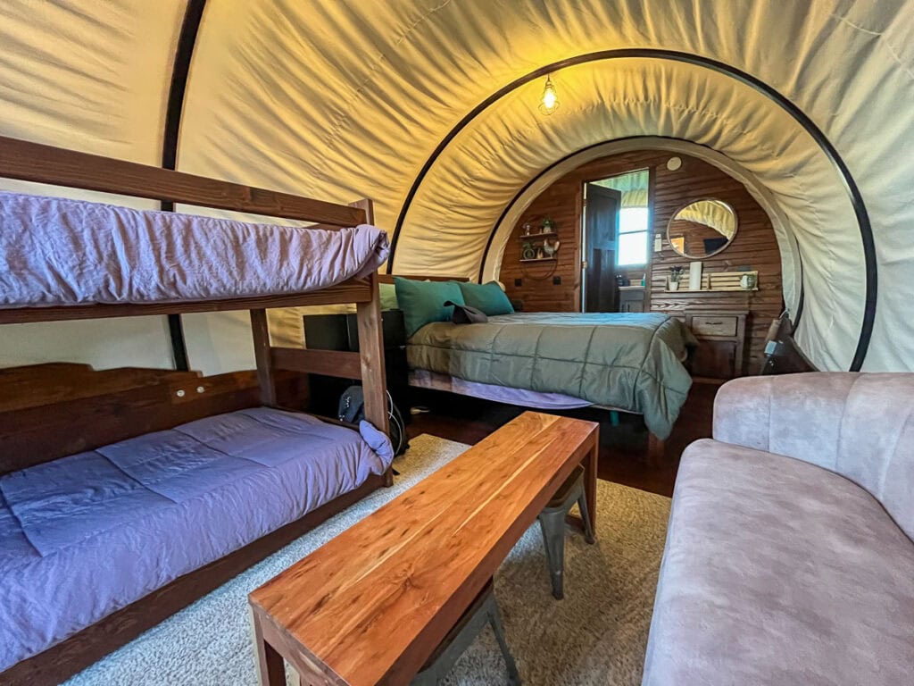 Interior of a cozy cabin, tent-like room with a bunk bed, queen-size bed, wooden bench, sofa, and small wooden table. Light enters from a doorway leading to a bathroom with mirror and sink—perfect for your next family trip.