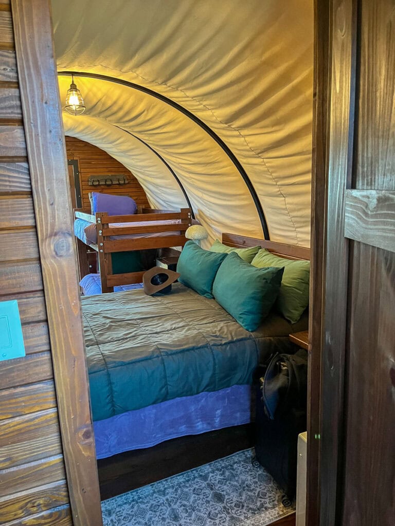 Perfect for a cozy cabin trip, this bedroom boasts a wooden interior, inviting bed with green pillows and a purple blanket, a wooden bunk bed in the background, curved canvas ceiling, lantern light, and a soft patterned rug.