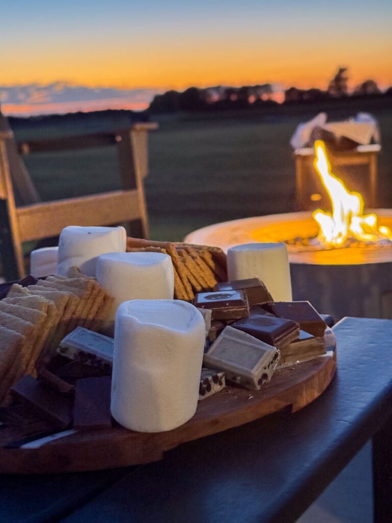 A close-up of a wooden tray with marshmallows, chocolate, and graham crackers by a fire pit at sunset—perfect for cozy cabin vibes. Two chairs and open fields in the background inspire great cabin ideas for your next family trip.