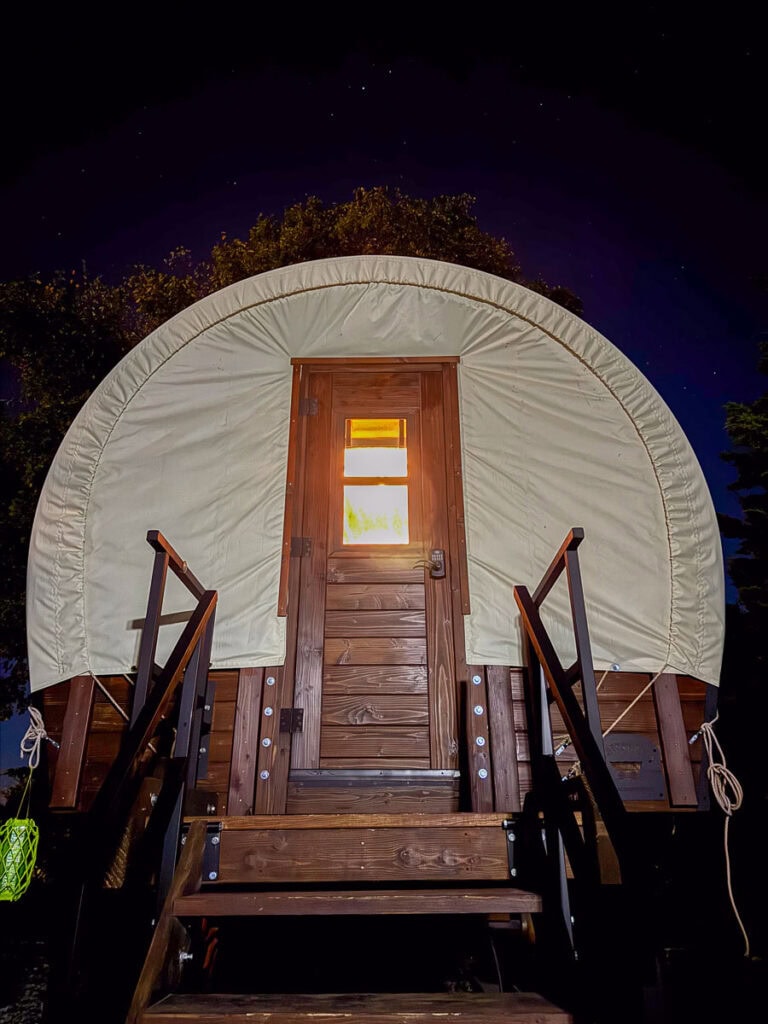 A lit covered wagon with wooden steps and a canvas roof is seen at night, illuminated from within—a unique twist on cozy cabin experiences, perfect for family getaway or cabin trip ideas under a starry sky.