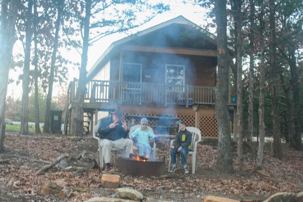 Three people sit in lawn chairs around a campfire in front of a cozy cabin surrounded by trees. Smoke rises from the fire as the group relaxes, enjoying nature and sharing ideas for their next family trip.