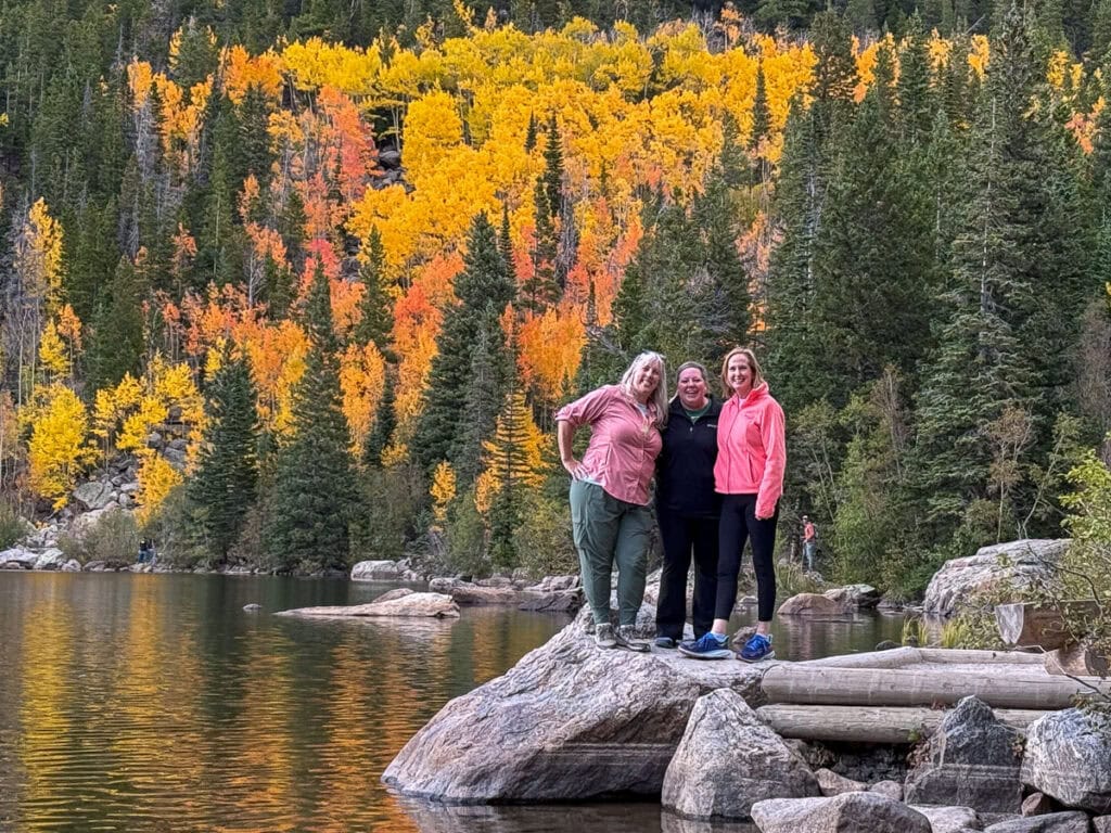Three women stand on a rock by a calm lake, smiling in front of a forested hillside with vibrant autumn foliage. Pine trees and rocky terrain surround them, creating the perfect backdrop for cozy cabin trip ideas.