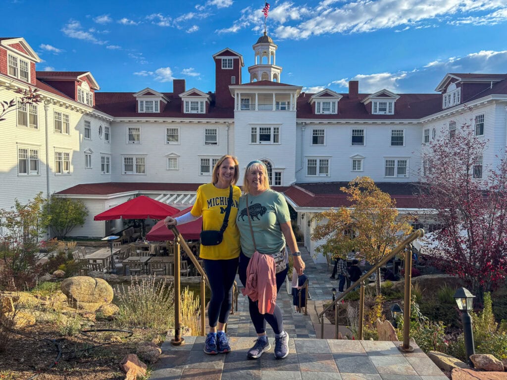 Two women stand smiling on outdoor steps in front of a large white historic hotel with a red roof, under a blue sky—a charming spot that inspires cozy cabin trip ideas, with bushes and patio furniture welcoming guests by the entrance.