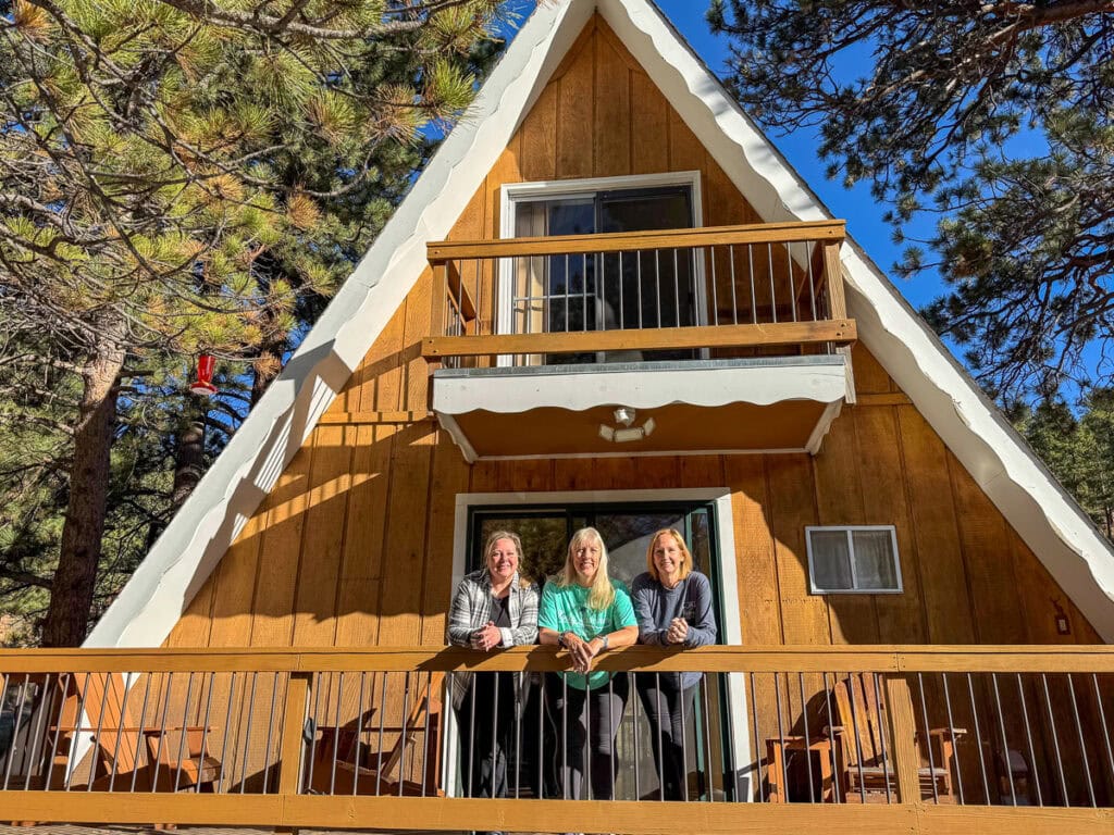 Three women stand and smile on the front deck of a brown A-frame cabin surrounded by tall pine trees—a perfect scene for those seeking cozy cabin trip ideas on a sunny day.