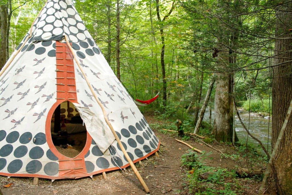 A white teepee with black circle patterns stands in a forest near a stream—perfect for cozy cabin trip ideas. The entrance is open, and a red hammock hangs between trees as sunlight filters through the green leaves.