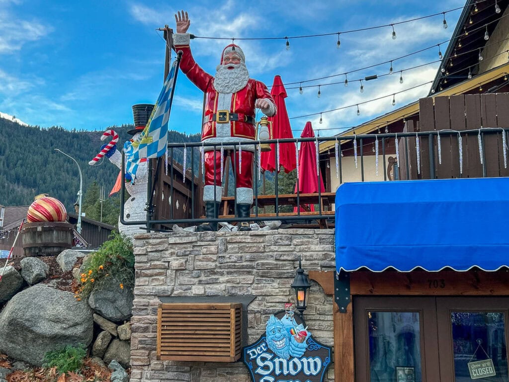 A large Santa Claus statue stands on a stone balcony, waving, outside a building decorated with Christmas lights—perfect inspiration for cozy cabin trip ideas. Mountains and a blue sky create a magical festive backdrop.