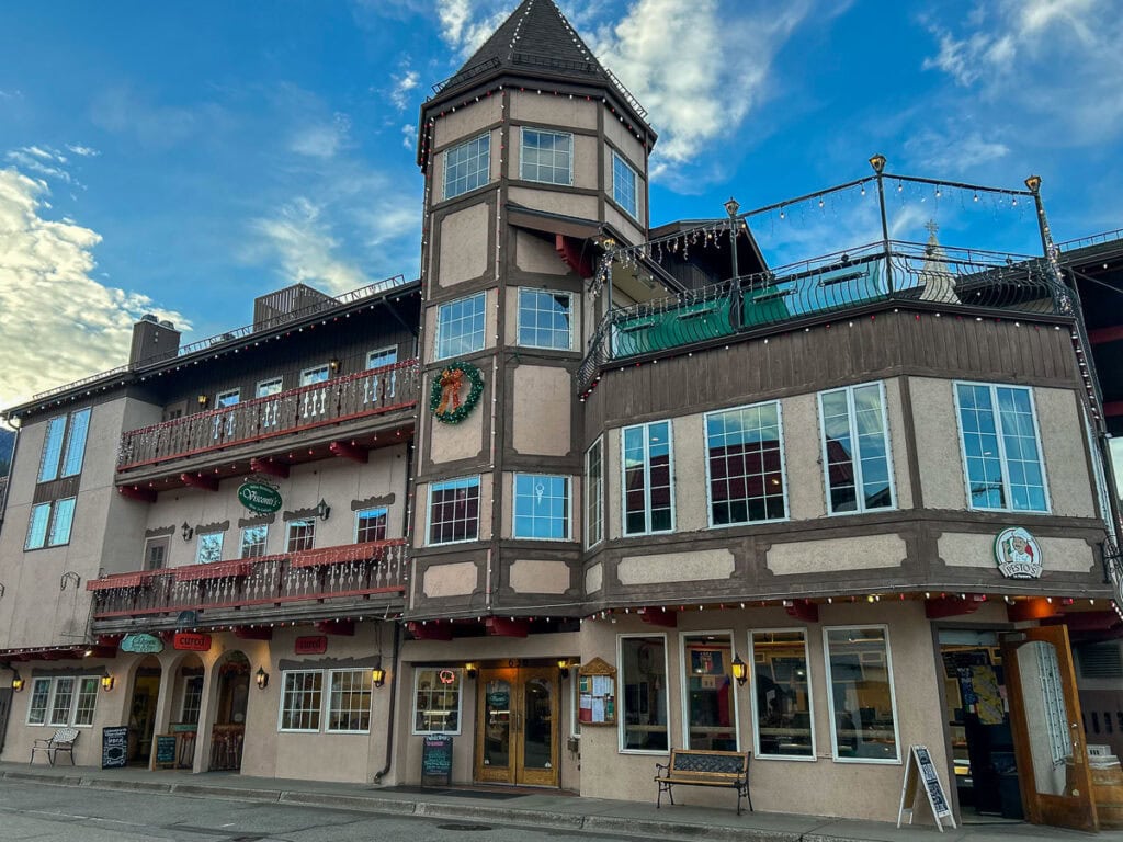 A three-story building with Bavarian-style architecture, featuring timber framing, balconies with red railings, a tower, and string lights—perfect inspiration for cozy cabin trip ideas. The sky is blue with scattered clouds.