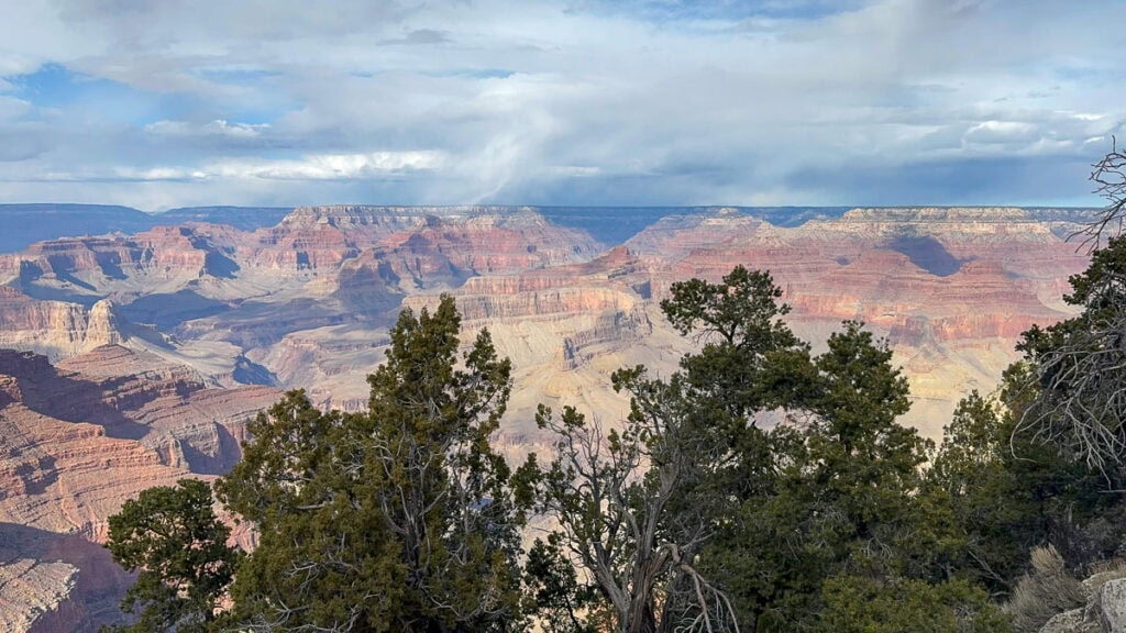 Scenic view of the Grand Canyon with layers of colorful rock formations, deep valleys, and tall green trees in the foreground under a partly cloudy sky—perfect inspiration for cozy cabin trip ideas.