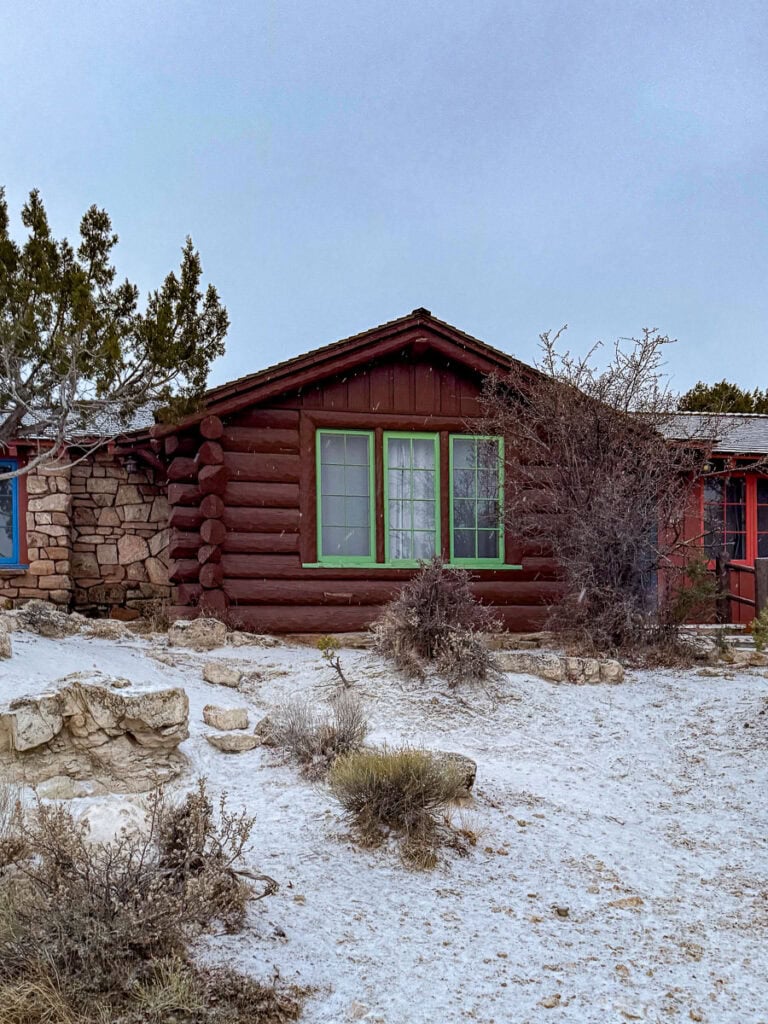 A rustic log cabin with green-trimmed windows sits on snowy ground, surrounded by shrubs and rocks under an overcast sky—perfect inspiration for cozy cabin trip ideas. Some trees are visible in the background.