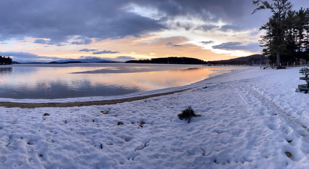 A snowy lakeshore at sunset, with calm water reflecting orange and purple hues in the sky—perfect for cozy cabin trip ideas. Pine trees line the right side, and distant hills are visible across the lake.
