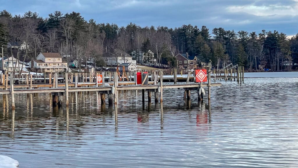 A wooden dock extends over calm water, with red signs posted along its length—perfect inspiration for cozy cabin trip ideas. Houses and trees line the distant shoreline under a partly cloudy sky.