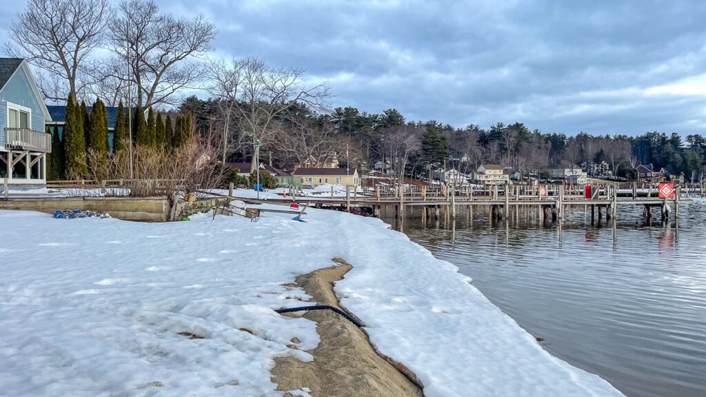 Snow covers the ground along a lakeshore, with docks extending into the water and houses in the background—offering cozy cabin trip ideas amid leafless trees under a cloudy sky.