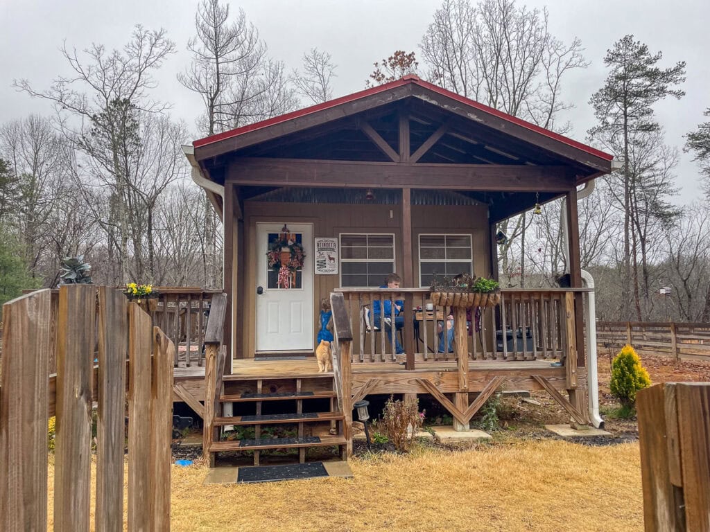 A cozy cabin with a covered porch sits in a yard with brown grass. Two people and a dog relax on the porch, surrounded by bare trees on a cloudy day—perfect for gathering cabin ideas for your next family trip.
