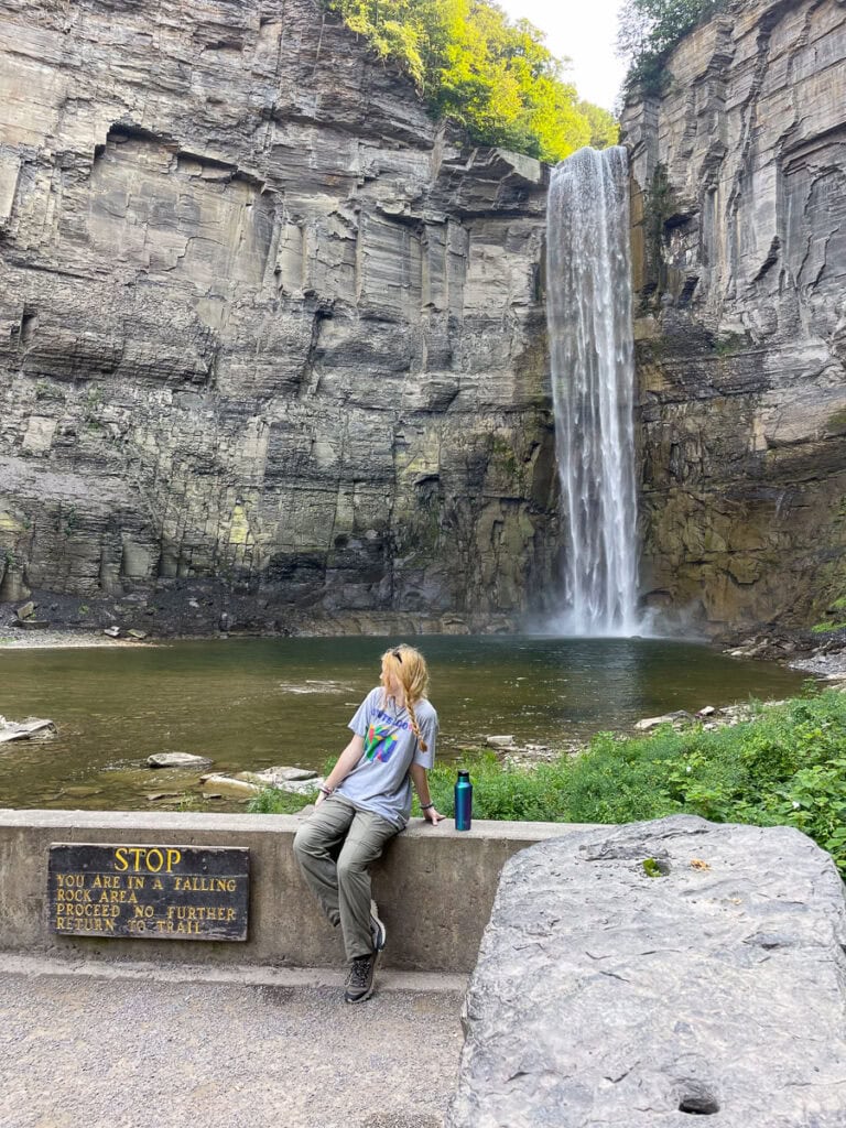 A person sits on a stone barrier near a sign warning of a falling hazard, facing a tall, narrow waterfall cascading down a rocky cliff into a pool—an inspiring view for your next cozy cabin trip ideas, all surrounded by lush greenery.