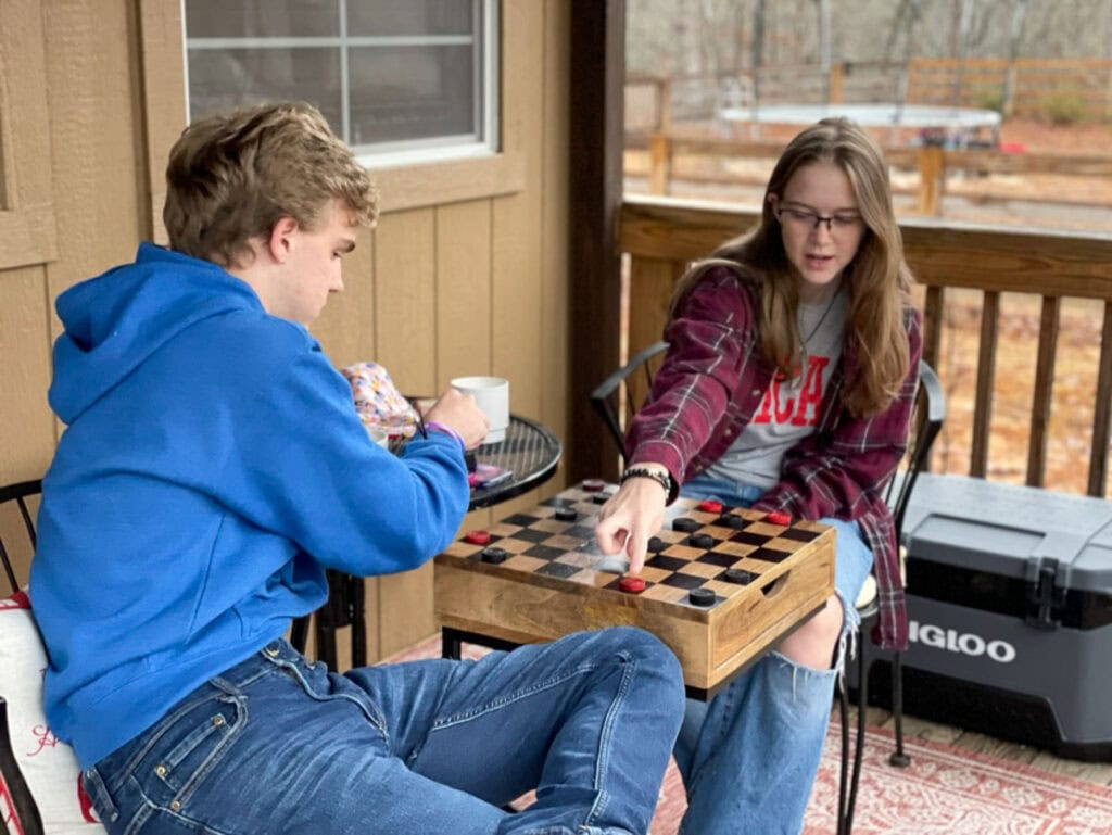 Two young people sit on a porch playing checkers during a cozy cabin vacation. The person on the left in a blue hoodie holds a mug, while the one on the right, in glasses and plaid shirt, moves a checker piece. An Igloo cooler sits in the background.