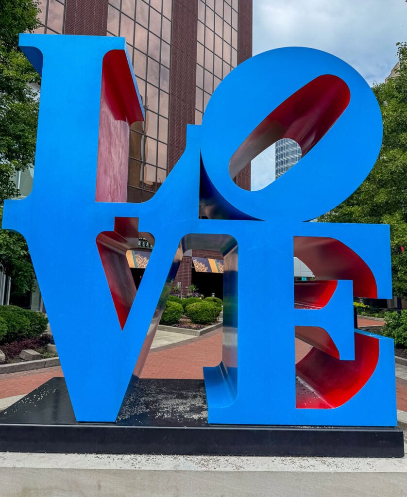 A large blue and red sculpture spells the word "LOVE," with the "O" tilted sideways. This iconic artwork is a must-see addition to your Grand Rapids itinerary, standing outdoors on a plaza with trees and a glass building in the background.