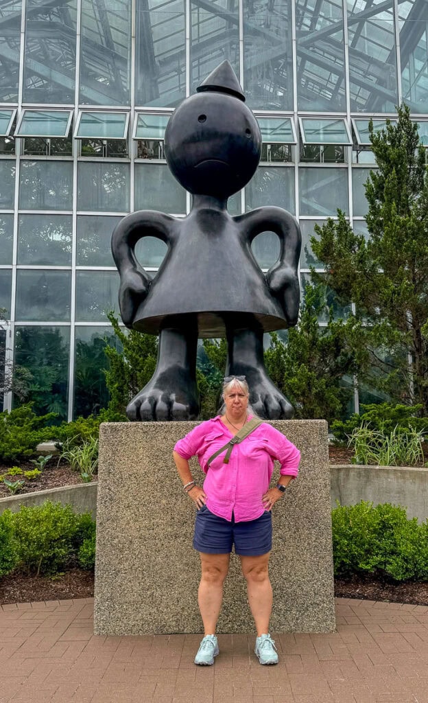 A woman in a pink shirt and navy shorts stands with hands on hips before a large black cartoonish statue, arms akimbo, outside a glass building—an ideal photo stop on any Grand Rapids itinerary.
