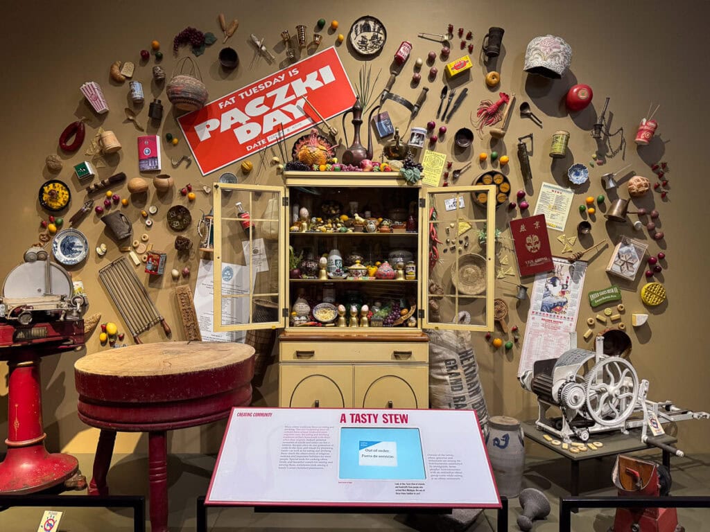 A vibrant museum display enriches any Grand Rapids itinerary, featuring a vintage kitchen scene with shelves of dishes and food, cooking tools, and Polish cultural items. A large “Paczki Day” sign hangs above, celebrating Fat Tuesday.