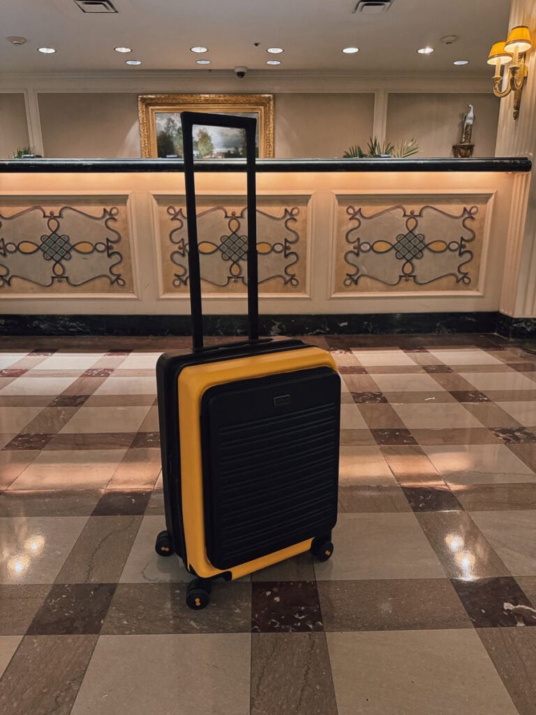 A yellow and black Level8 Carry-On hard shell suitcase with an extended handle stands on a checkered tile floor in front of an ornate hotel reception desk.