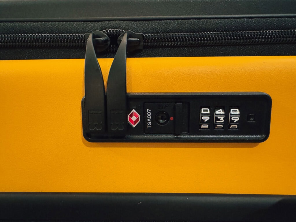 Close-up of a yellow Level8 Carry-On hard shell luggage with a black zipper and TSA lock featuring a combination dial and red diamond symbol. The lock is set to 0-0-0, perfect for any suitcase review.
