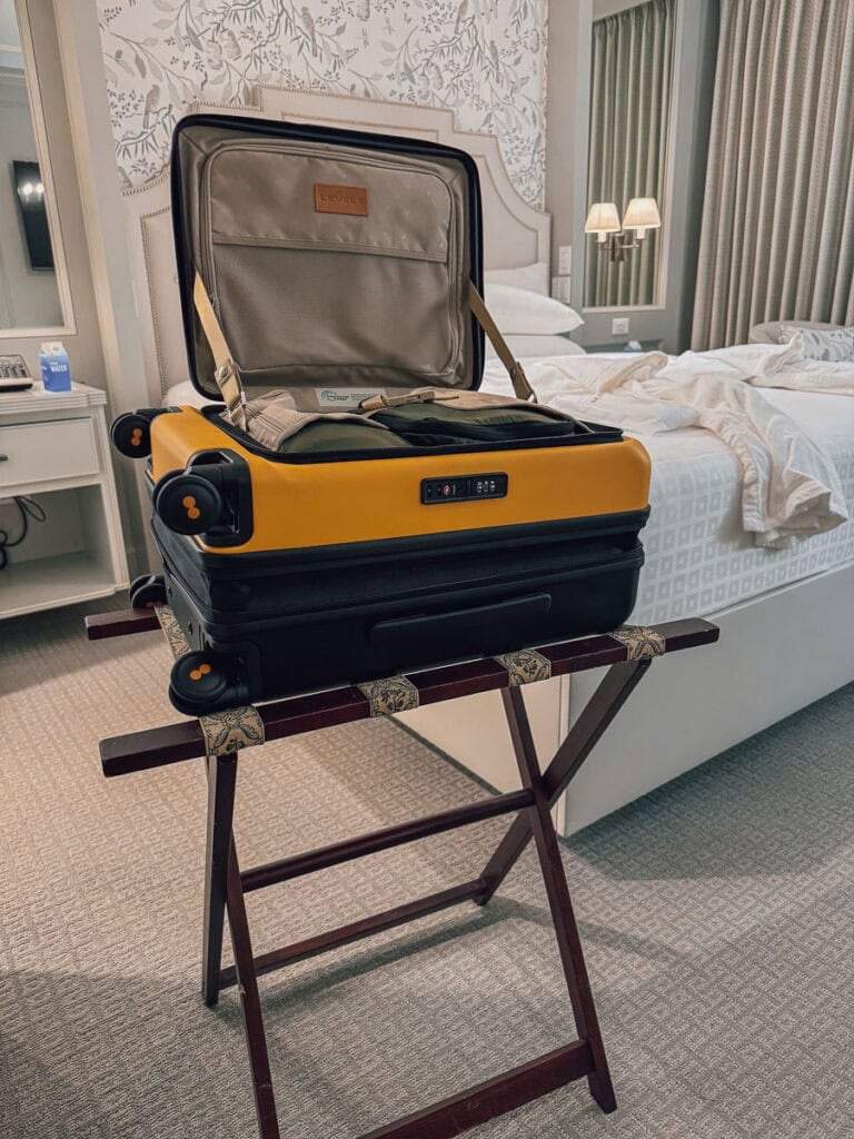 A yellow and black Level8 Carry-On hard shell luggage with an open lid sits on a wooden luggage stand in a neatly decorated hotel room with a made bed and wall lamps in the background.