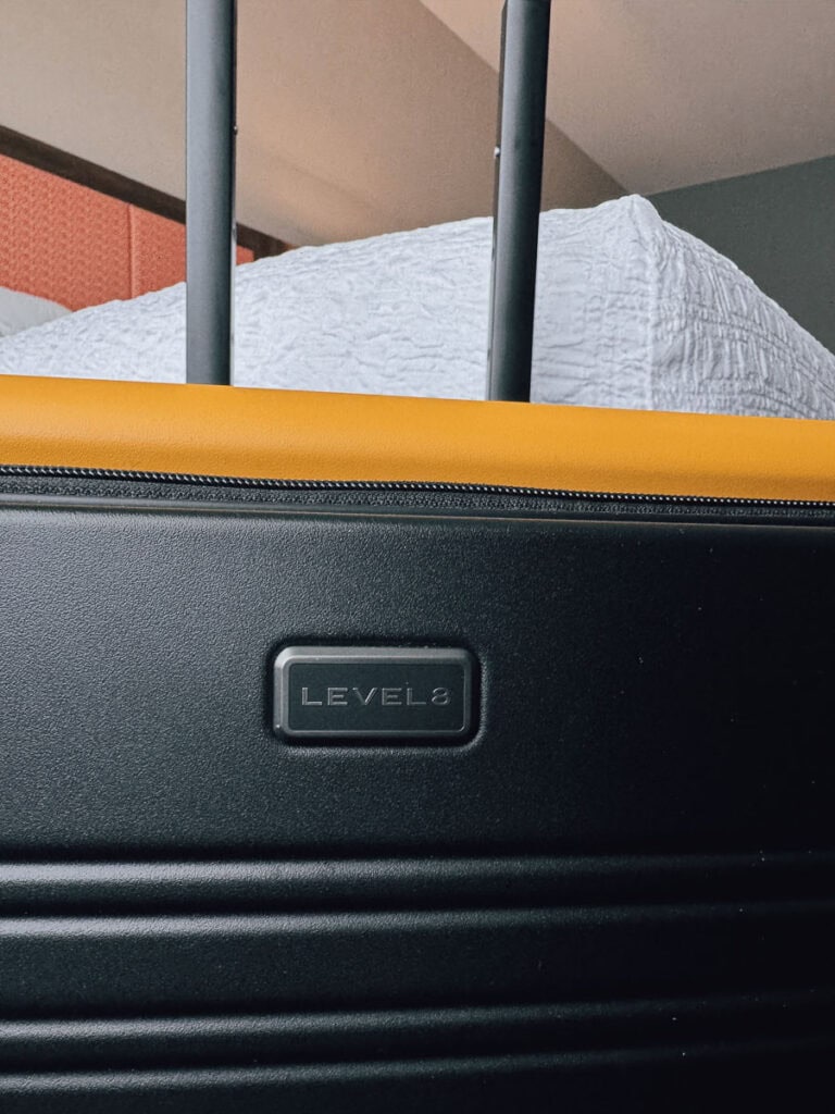 A close-up of a black LEVEL8 suitcase with a yellow zipper, showing the brand label and extended handles against a background with a white textured bedspread.