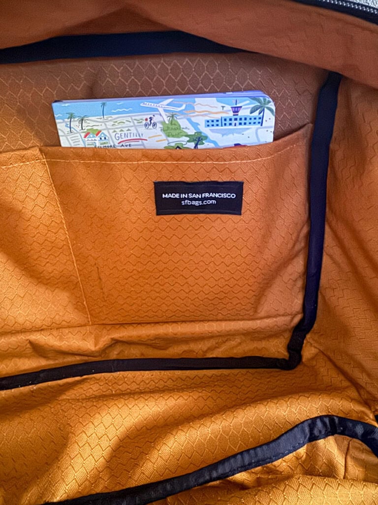 An open orange underseat bag with a pattern lining, showing a pocket containing a postcard with travel-themed illustrations. A label inside the pocket reads “MADE IN SAN FRANCISCO sfbag.com.” Perfect for frequent flyers.