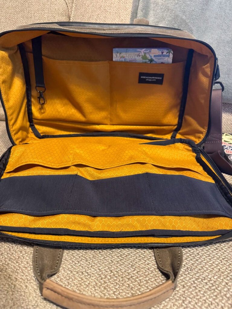 An open tan and yellow laptop-friendly underseat bag on a beige surface, showing organized interior pockets, including one holding a card and a folded paper, with a handle and visible zipper—perfect for frequent flyers. best laptop-friendly underseat bag.