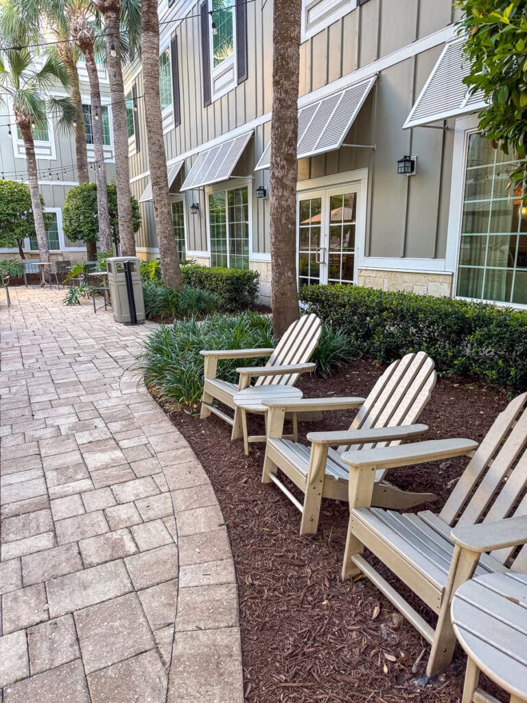 Three empty Adirondack chairs sit on mulch beside a paved walkway outside a modern building with large windows, white trim, palm trees, and neatly trimmed bushes—a tranquil spot that could easily be part of any relaxing New Smyrna Beach itinerary.