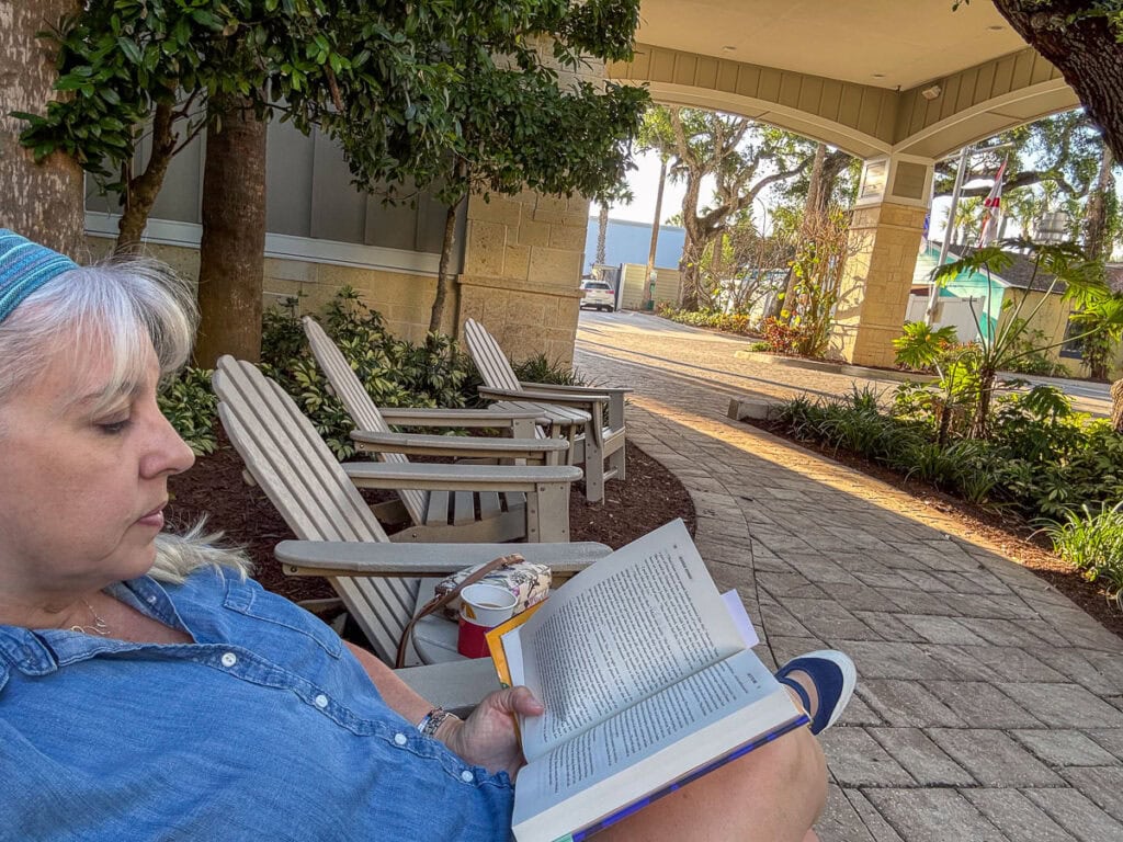A woman with gray hair in a blue shirt reads a book while sitting on an outdoor chair near a building, surrounded by trees and more empty chairs on a sunny day, perhaps planning her next new smyrna beach itinerary.